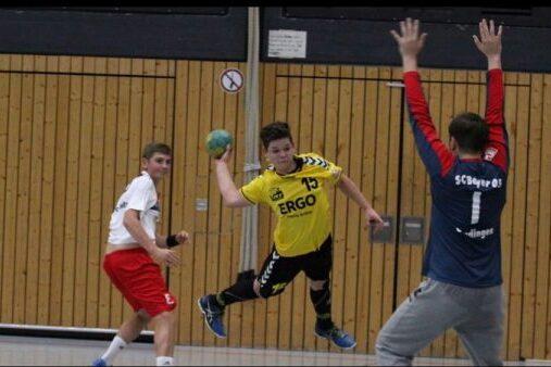 Handballspieler in gelb wirft auf das Tor, w&auml;hrend zwei Gegner in wei&szlig; und rot defending. Sporthalle mit Holzpaneelen im Hintergrund.