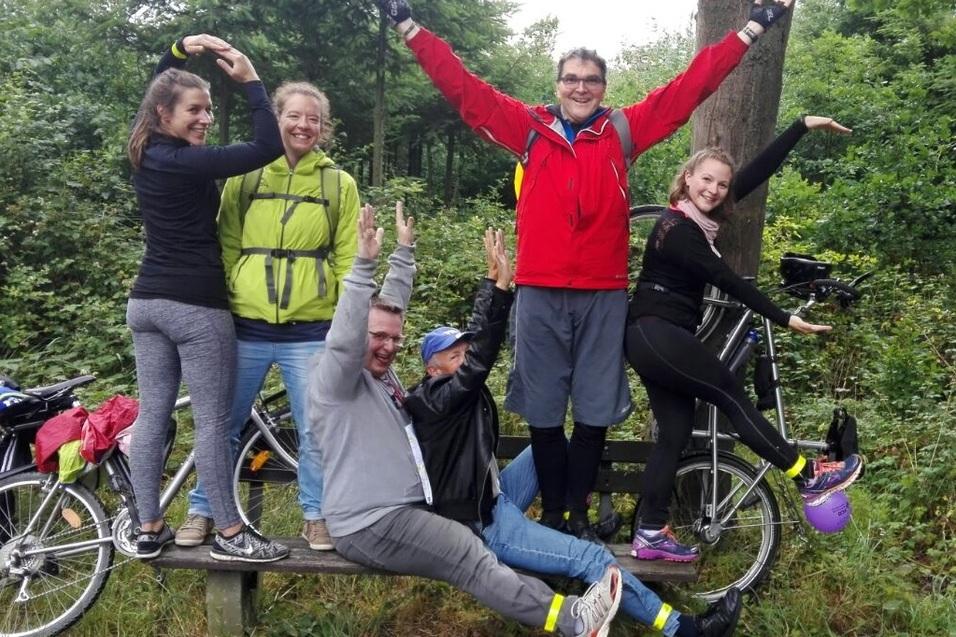 Gruppe von sechs Personen, die auf einem Fahrrad und einer Holzbank posieren, umgeben von gr&uuml;ner Waldlandschaft.