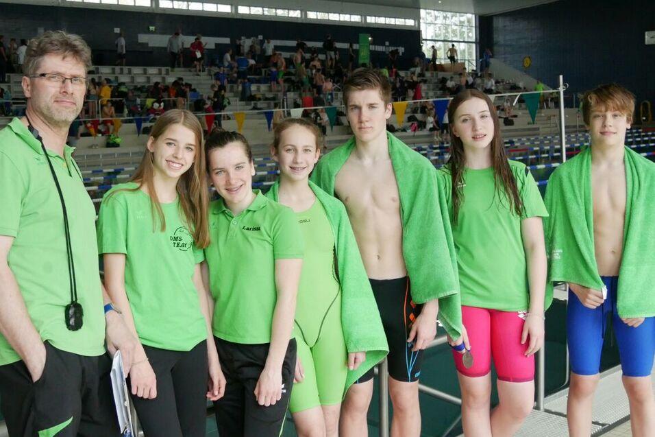 Sechs Schwimmer und ein Trainer stehen am Beckenrand, alle tragen gr&uuml;ne Shirts; einige mit Handt&uuml;chern, im Hintergrund Zuschauer.