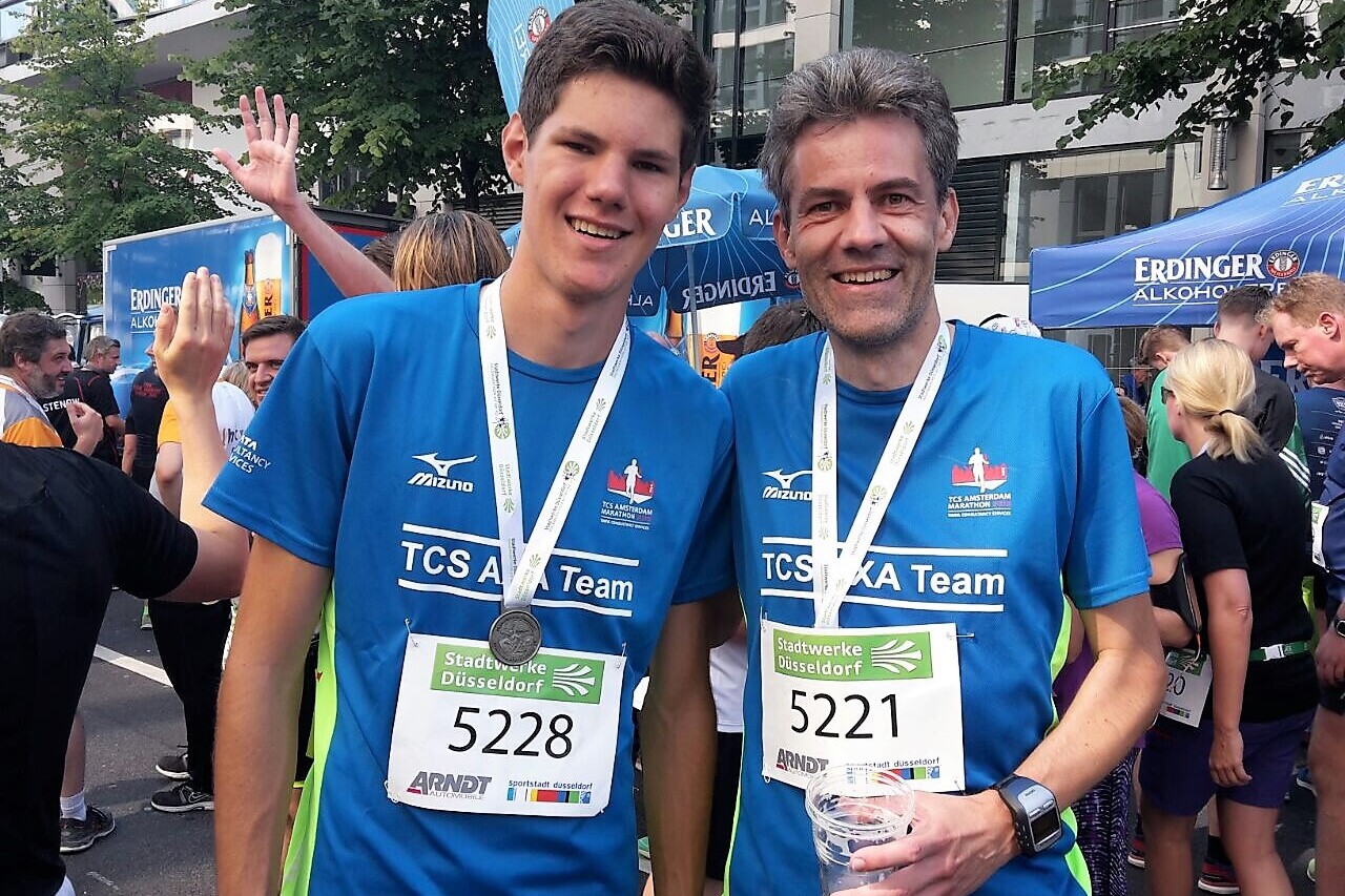 Zwei L&auml;ufer in blauen T-Shirts mit Medaillen stehen l&auml;chelnd vor einem Rennveranstaltungs-Hintergrund.