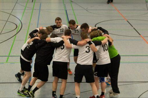 Eine Gruppe von Handballspielern huddelt sich in einer Sporthalle vor dem Spiel. Einige tragen Trikots mit Nummern.
