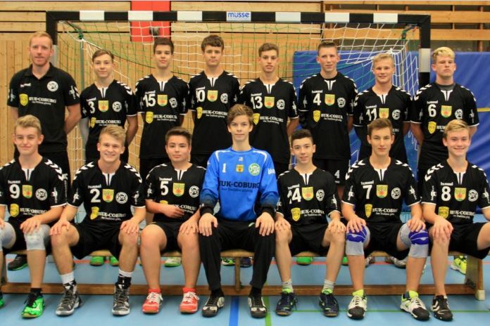 Mannschaftsbild einer Jugend-Handballmannschaft in schwarzen Trikots, sitzend und stehend vor einem Handballtor.