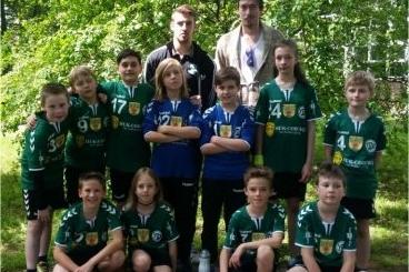 Gruppenfoto von zwei Trainern und einer Jugendhandballmannschaft in Sportkleidung vor grüner Naturkulisse.