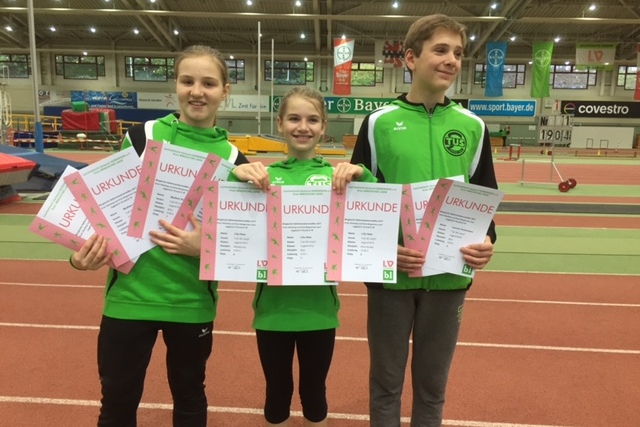 Drei Kinder in gr&uuml;nen Sportoutfits halten Urkunden in einer Sporthalle mit Laufbahn im Hintergrund.