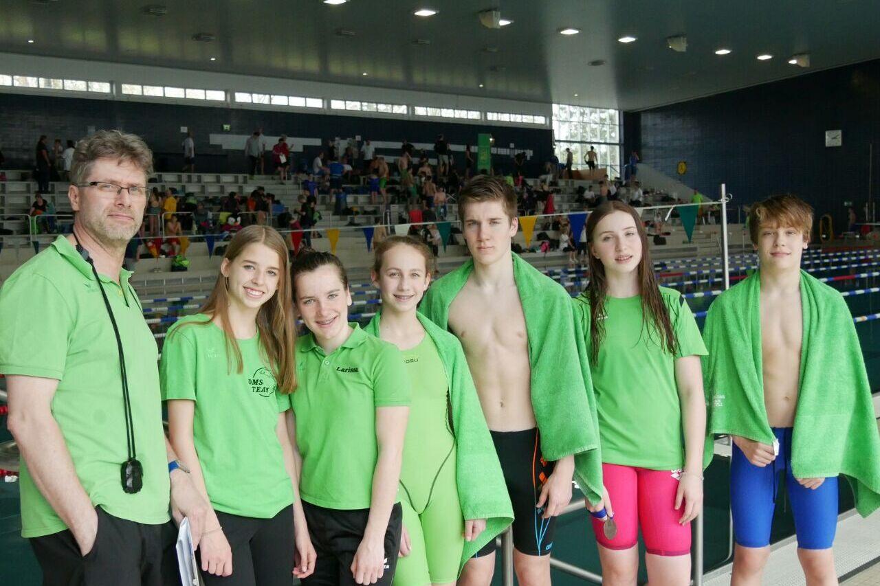 Sechs Schwimmer und ein Trainer stehen am Beckenrand, alle tragen grüne Shirts; einige mit Handtüchern, im Hintergrund Zuschauer.