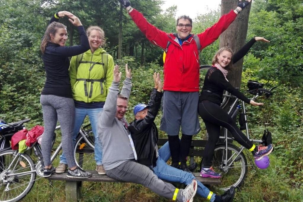 Gruppe von sechs Personen, die auf einem Fahrrad und einer Holzbank posieren, umgeben von grüner Waldlandschaft.