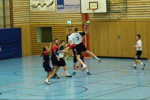 Ein Handballspieler springt hoch, um zu werfen, w&auml;hrend Gegner ihn verteidigen. Halle mit blauem Boden und Holzagen.
