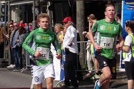 Zwei junge L&auml;ufer in Sportbekleidung rennen bei einer Laufveranstaltung, Zuschauer stehen am Rand.