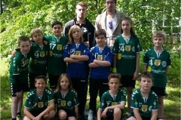 Gruppenfoto von zwei Trainern und einer Jugendhandballmannschaft in Sportkleidung vor gr&uuml;ner Naturkulisse.