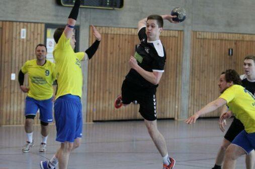 Handballspieler springt mit Ball in der Hand, w&auml;hrend ein Gegner versucht, ihn zu blockieren. Weitere Spieler im Hintergrund.