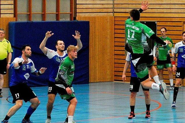Spielszene im Handball mit einem Spieler in gr&uuml;n, der springt, w&auml;hrend andere Spieler in blauen Trikots abwehren.