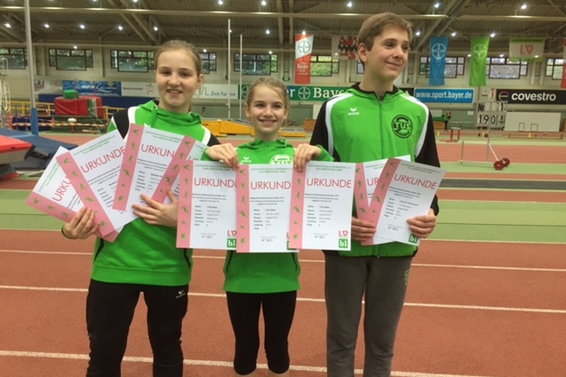 Drei Kinder in gr&uuml;nen Sportoutfits halten Urkunden in einer Sporthalle mit Laufbahn im Hintergrund.