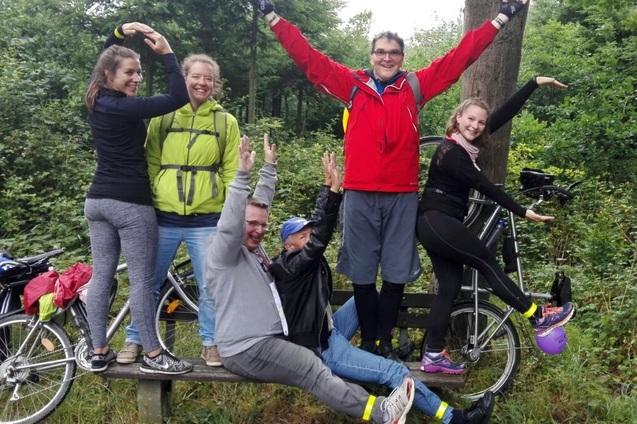 Gruppe von sechs Personen, die auf einem Fahrrad und einer Holzbank posieren, umgeben von gr&uuml;ner Waldlandschaft.