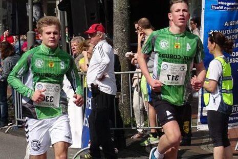 Zwei junge L&auml;ufer in Sportbekleidung rennen bei einer Laufveranstaltung, Zuschauer stehen am Rand.