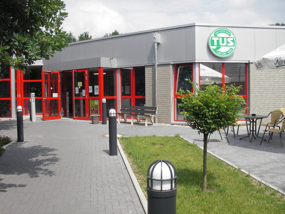 Eingang eines modernen Geb&auml;udes mit roten Fenstern und einem Logo, umgeben von Rasen und Terrassenm&ouml;beln.