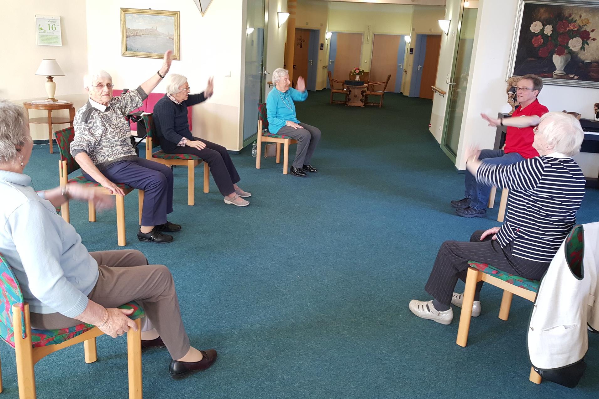 Gruppe älterer Menschen sitzt auf Stühlen und führt gemeinschaftliche Bewegungsübungen in einem hellen Raum durch.