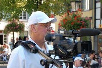 Mann mit wei&szlig;em Hut filmt mit einem Camcorder in einer belebten Innenstadt, umgeben von B&auml;umen und Menschen.