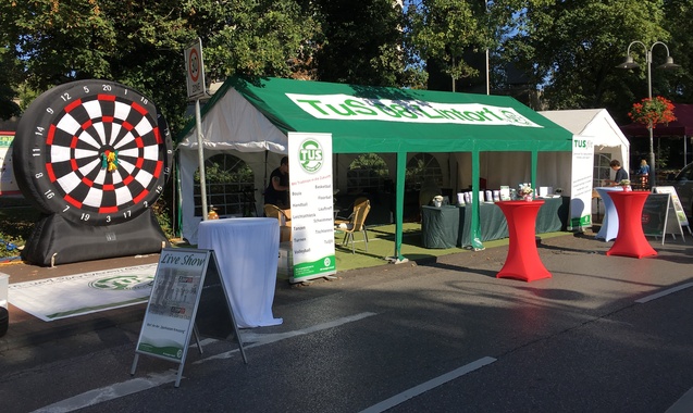 Stand mit einem Dartboard und einem Zelt, daneben mehrere Stehtische auf einer Veranstaltung im Freien.