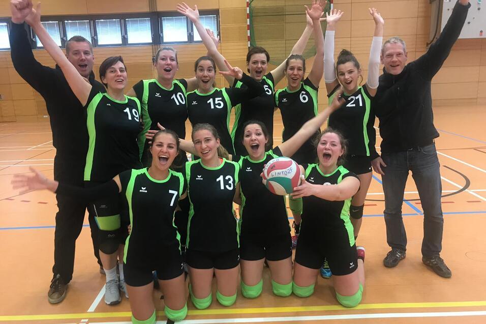 Freudige Volleyballmannschaft posiert mit einem Ball in der Mitte, umgeben von jubelnden Mitspielern in einheitlichen Trikots.