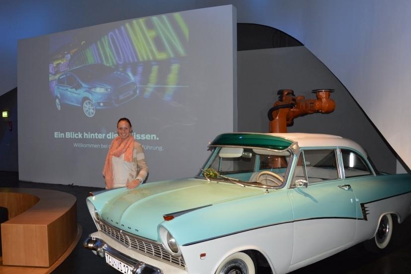 Frau mit rosa Schal posiert neben einem klassischen blauen Auto vor einer digitalen Pr&auml;sentation im Hintergrund.