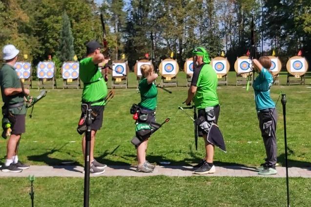 Sechs Bogensch&uuml;tzen in gr&uuml;nen Shirts zielen auf farbige Zielscheiben im Freien.