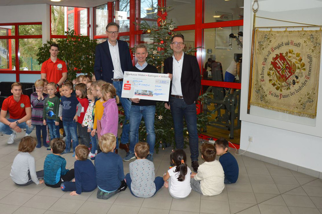 Gruppierung von Kindern und Erwachsenen in einem Raum, w&auml;hrend ein Scheck &uuml;berreicht wird, dekoriert mit einem Weihnachtsbaum.