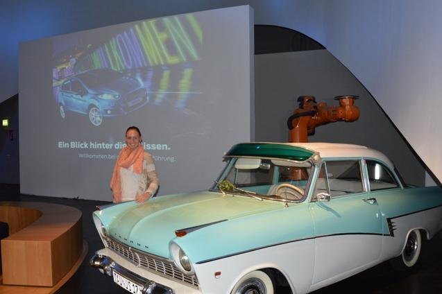 Frau mit rosa Schal posiert neben einem klassischen blauen Auto vor einer digitalen Pr&auml;sentation im Hintergrund.