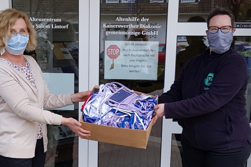 Zwei Frauen in Masken &uuml;bergeben einen Karton mit Masken vor einem Altenzentrum. Hintergrund zeigt ein Stoppschild.