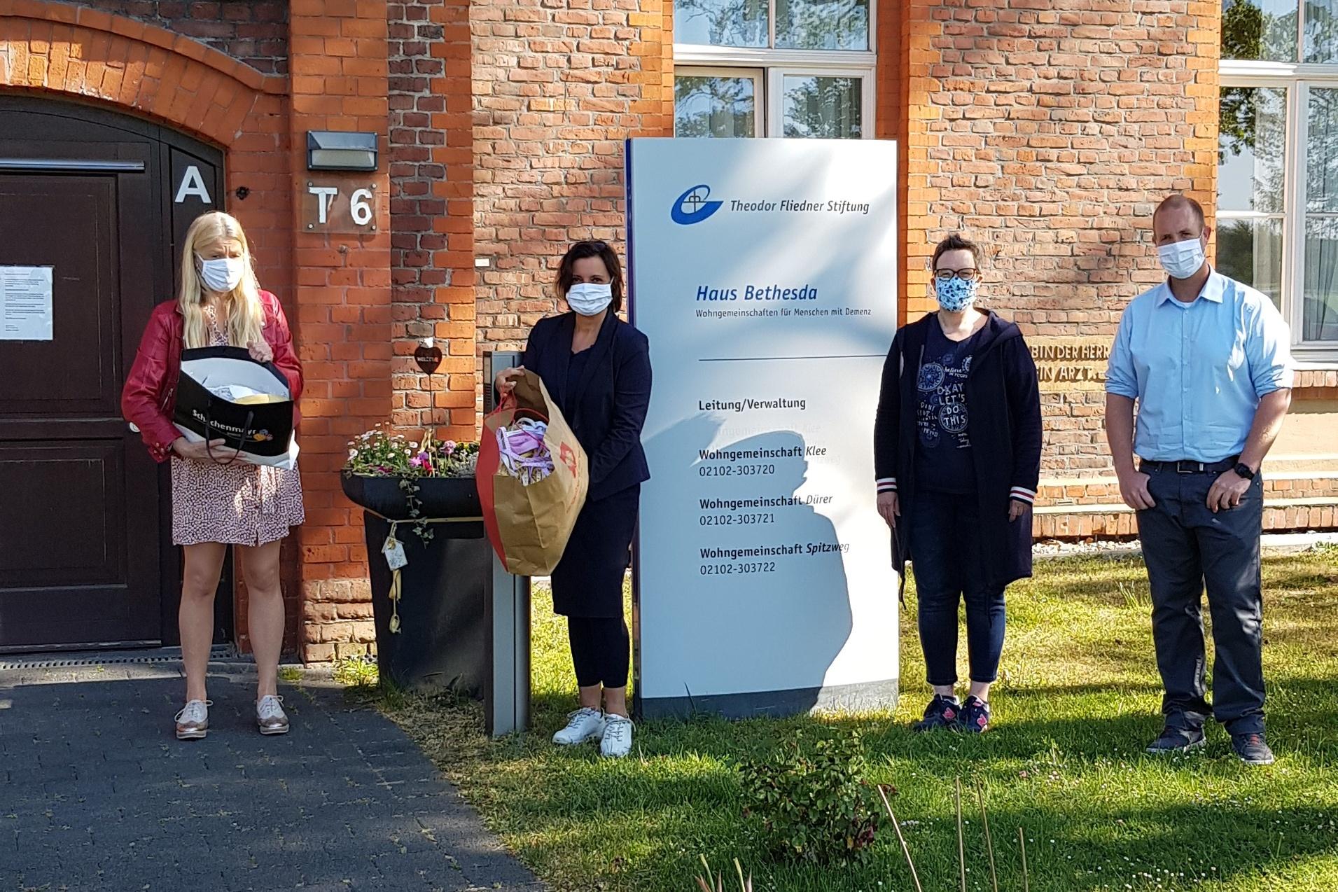 Vier Personen mit Mund-Nasen-Schutz stehen vor einem Backsteinbau, neben einem Schild mit der Aufschrift „Haus Bethesda“.