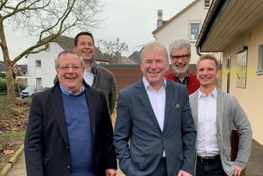 F&uuml;nf M&auml;nner stehen l&auml;chelnd auf einem Gehweg vor einem Wohnhaus, umgeben von B&auml;umen und einer grauen Hauswand.