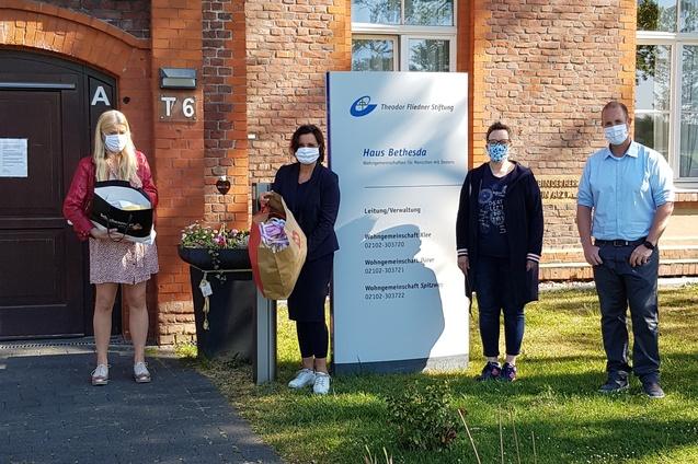 Vier Personen mit Mund-Nasen-Schutz stehen vor einem Backsteinbau, neben einem Schild mit der Aufschrift &bdquo;Haus Bethesda&ldquo;.