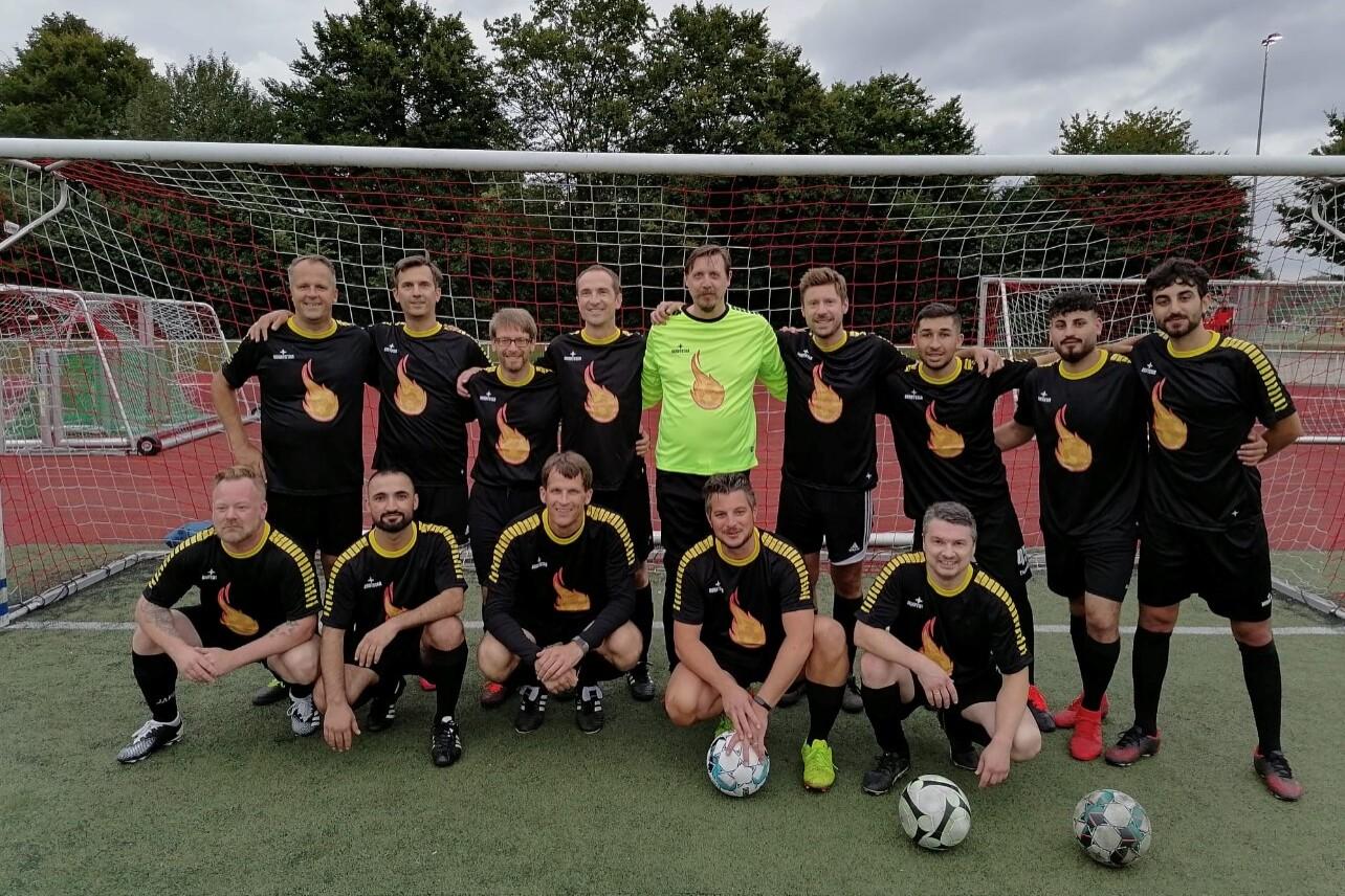 Gruppe von 14 Fu&szlig;ballspielern in schwarzen Trikots mit Flammenmotiv auf Sportplatz vor einem Tor.