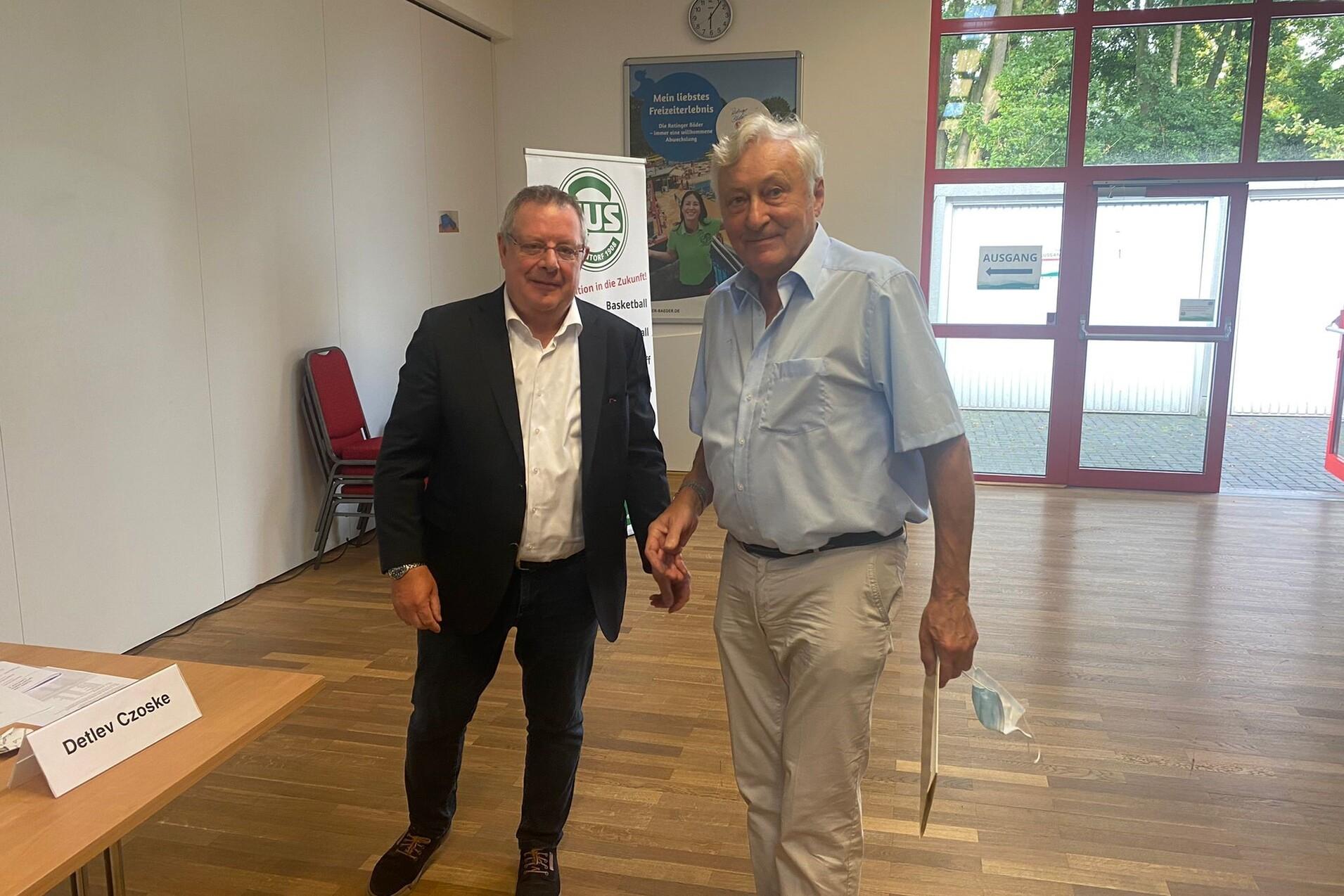 Zwei Männer stehen lächelnd nebeneinander in einem Raum mit Holzfußboden und Fensterfront.