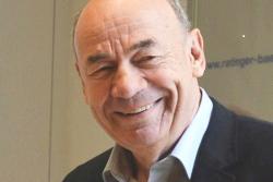 L&auml;chelnder Mann mit Glatze und Jacket, steht vor einer hellen Wand.