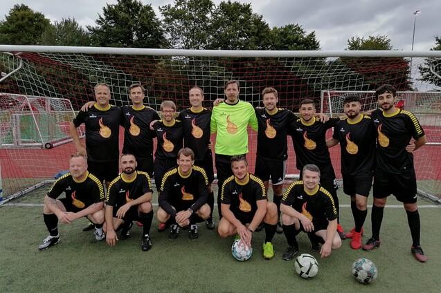 Gruppe von 14 Fu&szlig;ballspielern in schwarzen Trikots mit Flammenmotiv auf Sportplatz vor einem Tor.