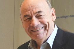 L&auml;chelnder Mann mit Glatze und Jacket, steht vor einer hellen Wand.