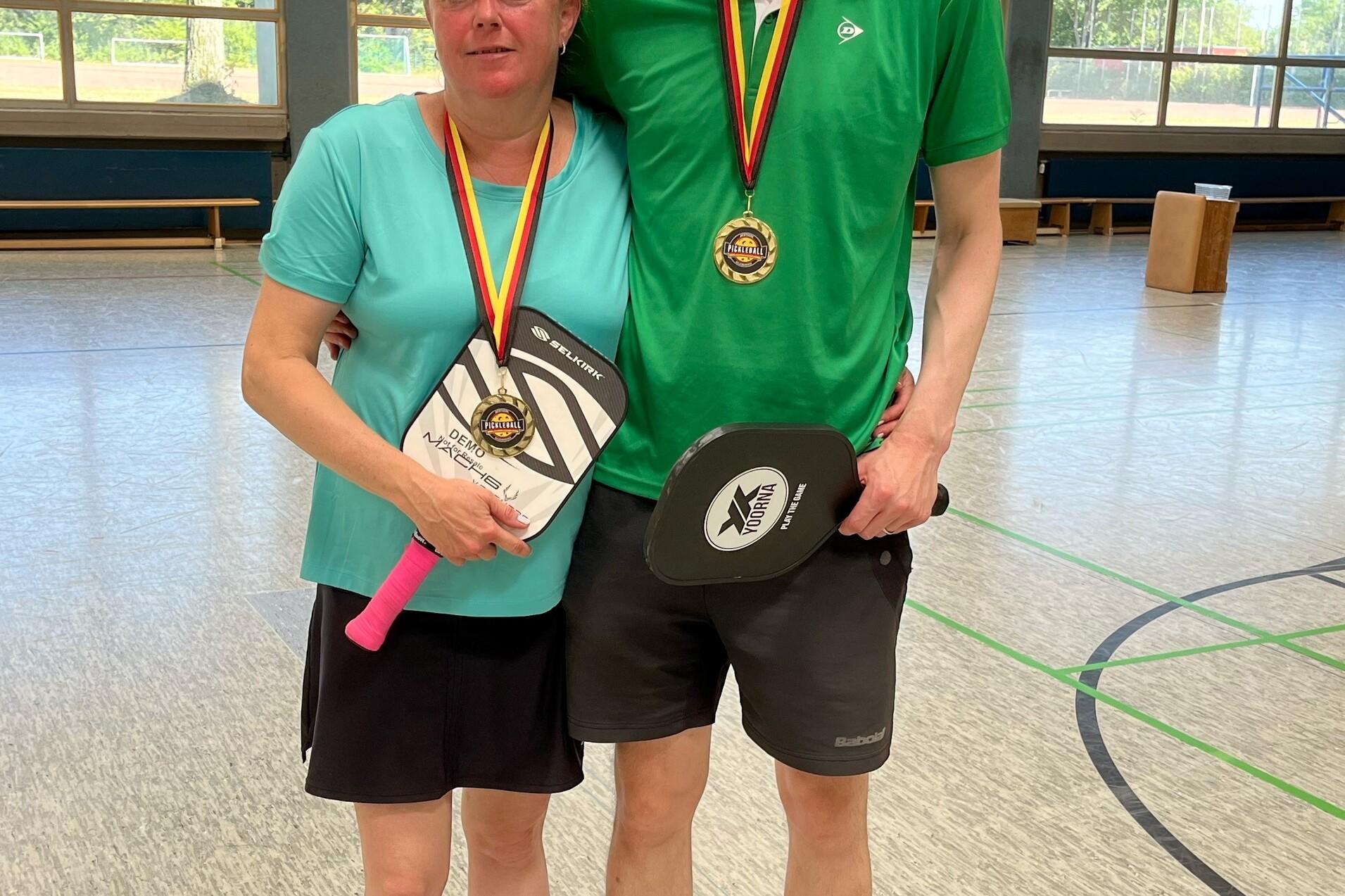Paar mit Medaillen und Pokalen in Sportkleidung steht in einer Turnhalle. Fenster mit Tageslicht im Hintergrund.