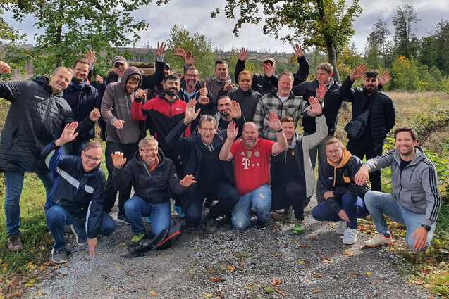 Gruppennfoto von 22 M&auml;nnern, die fr&ouml;hlich auf einem Kiesweg in der Natur posieren, umgeben von B&auml;umen und Wiesen.