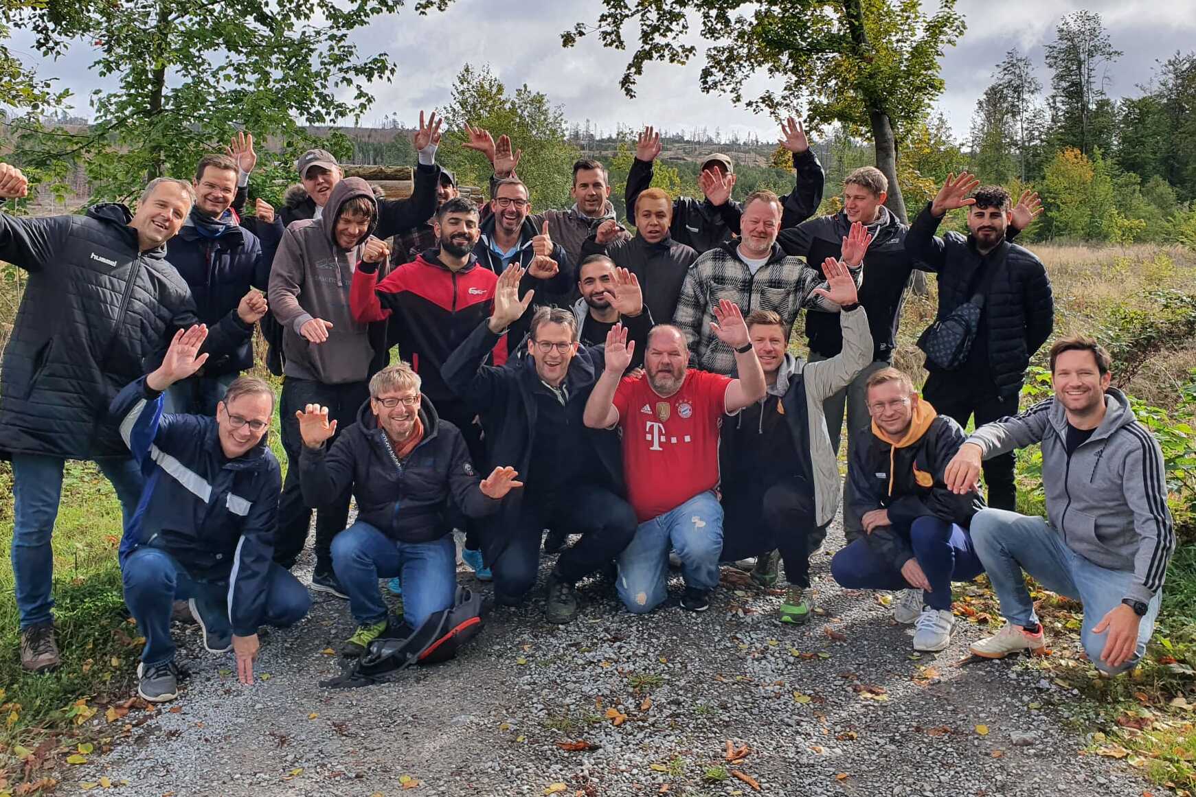 Gruppennfoto von 22 M&auml;nnern, die fr&ouml;hlich auf einem Kiesweg in der Natur posieren, umgeben von B&auml;umen und Wiesen.