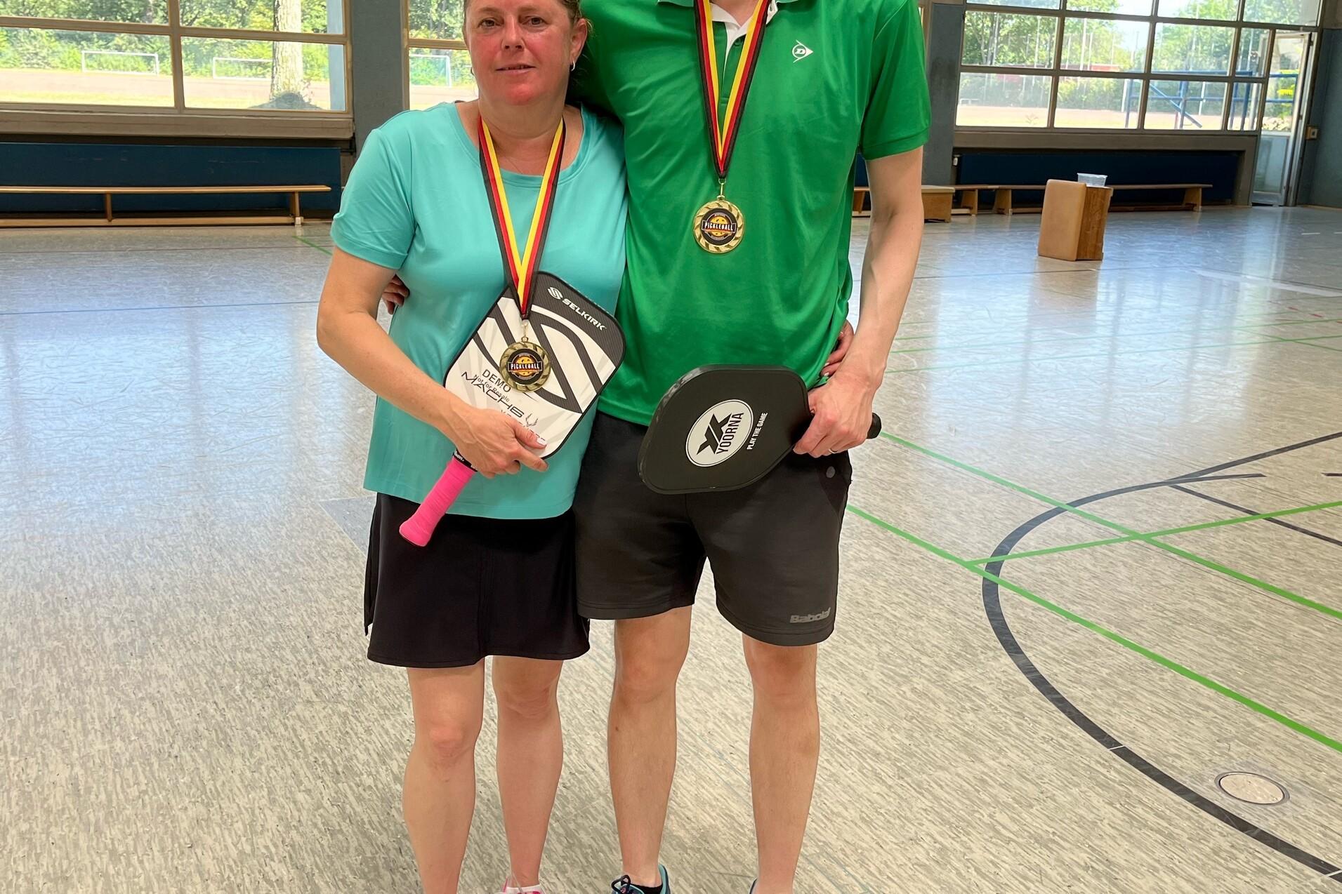 Paar mit Medaillen und Pokalen in Sportkleidung steht in einer Turnhalle. Fenster mit Tageslicht im Hintergrund.