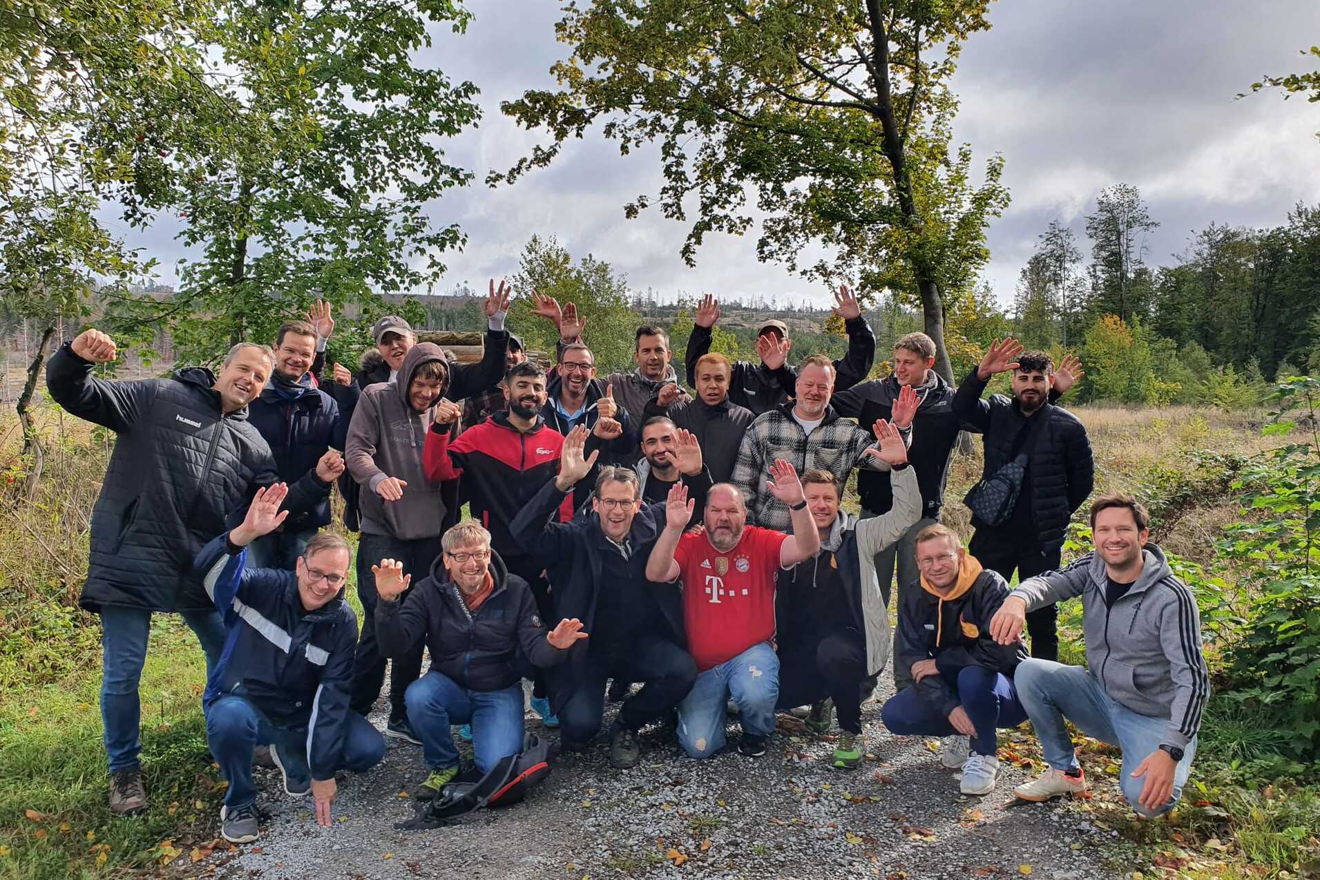 Gruppennfoto von 22 M&auml;nnern, die fr&ouml;hlich auf einem Kiesweg in der Natur posieren, umgeben von B&auml;umen und Wiesen.