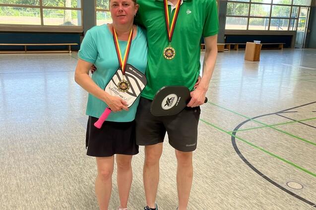 Paar mit Medaillen und Pokalen in Sportkleidung steht in einer Turnhalle. Fenster mit Tageslicht im Hintergrund.