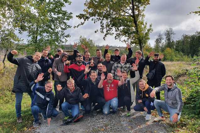 Gruppennfoto von 22 M&auml;nnern, die fr&ouml;hlich auf einem Kiesweg in der Natur posieren, umgeben von B&auml;umen und Wiesen.