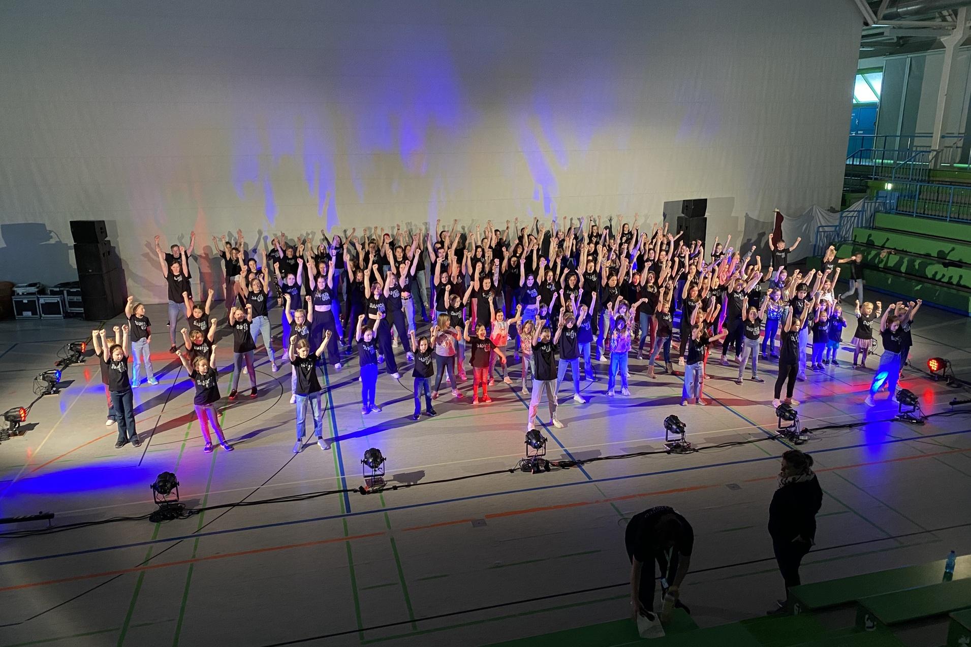 Große Gruppe von Tänzern in schwarzen Outfits mit erhobenen Händen auf einer beleuchteten Bühne in einer Sporthalle.