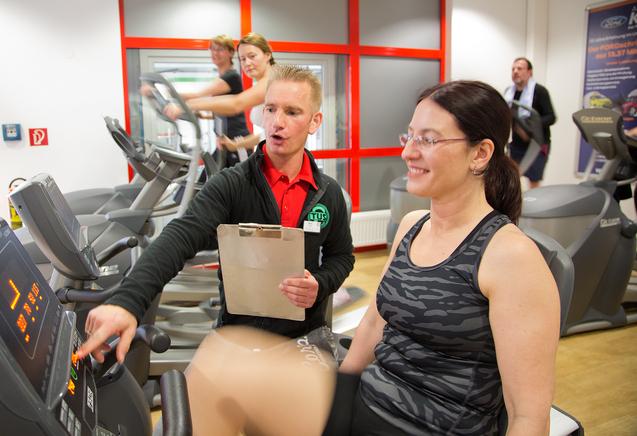 Trainer erkl&auml;rt einer Frau auf einem Crosstrainer die Einstellungen; im Hintergrund weitere Personen beim Training.