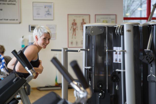 Junge Frau mit kurzen blonden Haaren l&auml;chelt, w&auml;hrend sie sich an einem Fitnessger&auml;t in einem Gym orientiert.