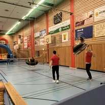 Zwei Sportler in roten Shirts trainieren mit einem Volleyball im Hallensportzentrum. Bretter und Werbebanner umgeben sie.