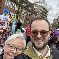 Selbstportr&auml;t eines Mannes und einer &auml;lteren Frau, umgeben von Personen in Kost&uuml;men w&auml;hrend einer Feier.