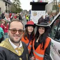 Drei Personen in leuchtenden Westen posieren vor einem Umzug mit bunten Festteilnehmern im Hintergrund.