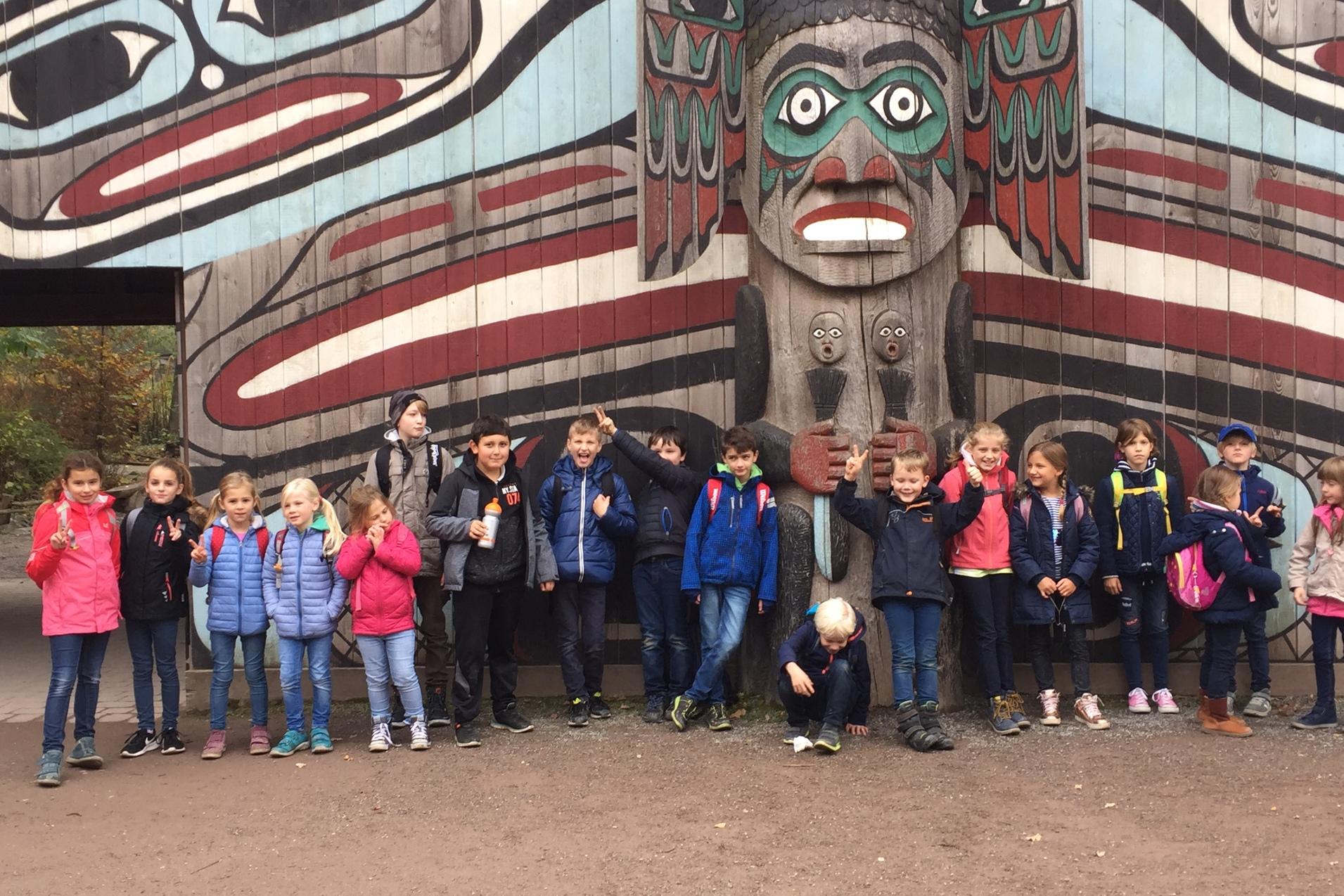 Gruppe von Kindern vor einer kunstvoll bemalten Totempfahlwand im Freien, l&auml;chelnd und teils posierend.